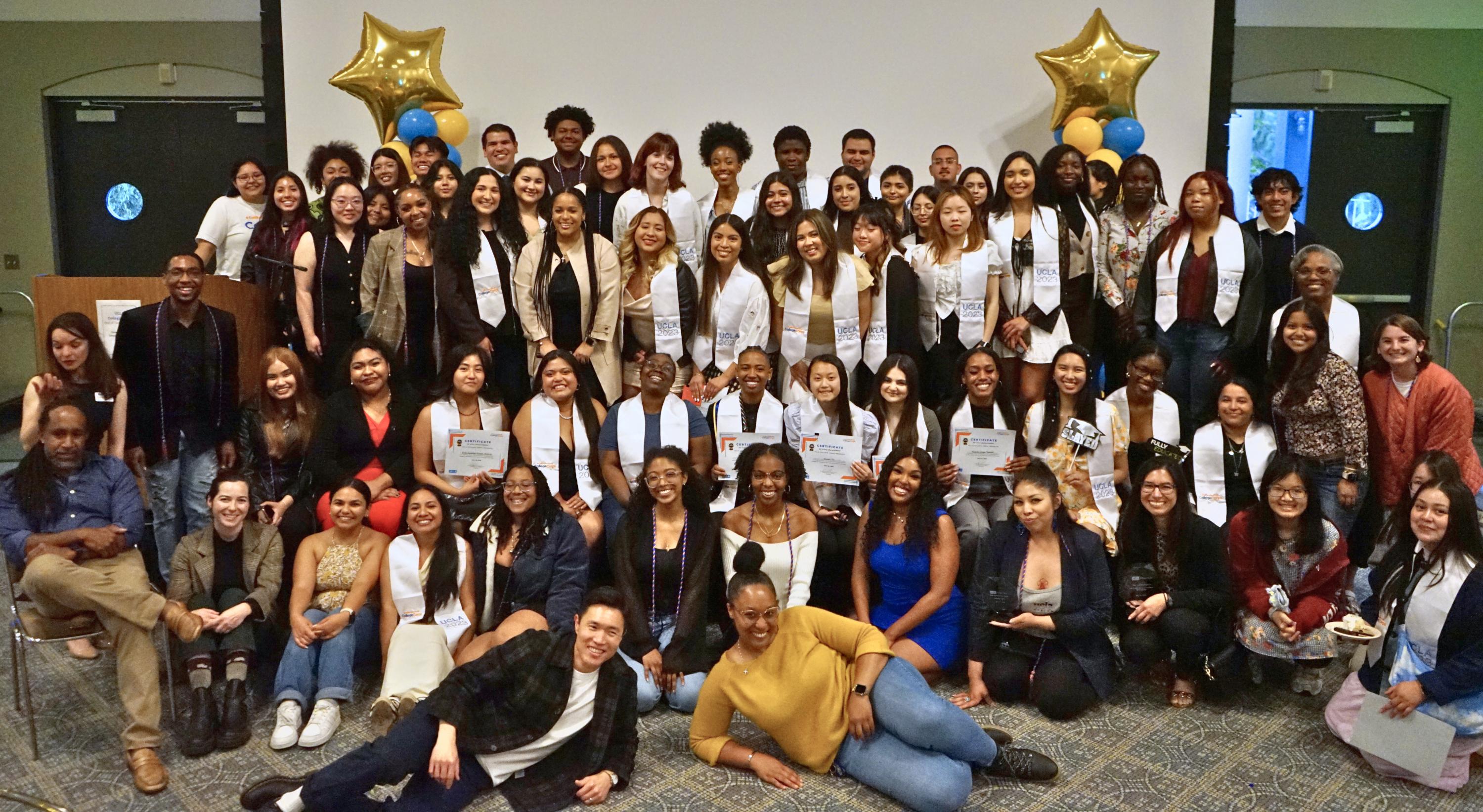 A large group of multicultural students and staff smiles for a photo in front of a large projection screen and yellow balloons shaped like stars.