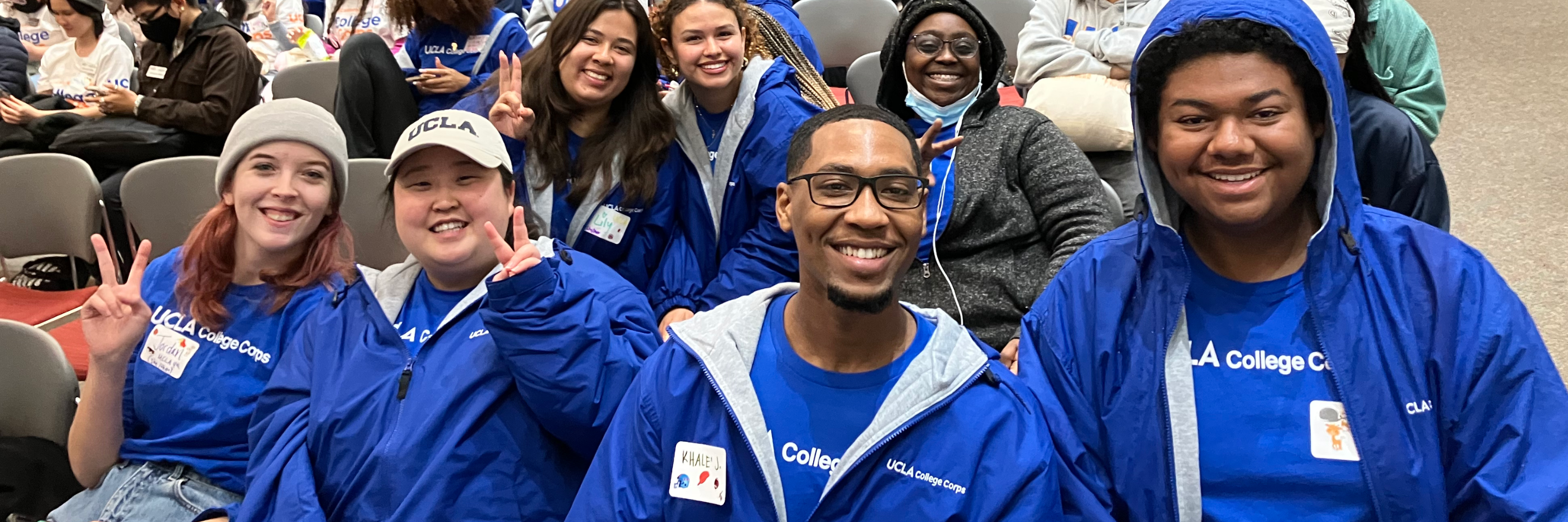 A group of students, 2 male and 5 female, in blue College Corp jackets smile for a photo